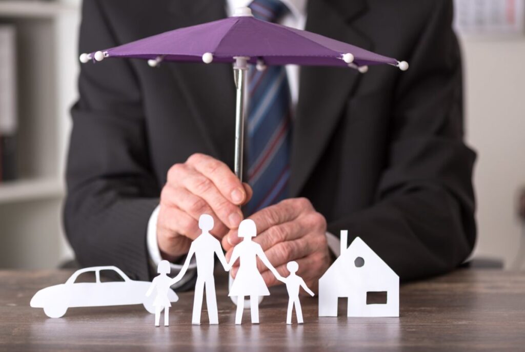 A businessman holding a small umbrella over paper cutouts of a family, car, and house, symbolizing personalized coverage for home, auto, and family insurance needs.